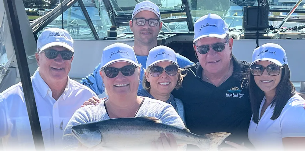 A group of six charter guests in matching caps share a salmon on deck